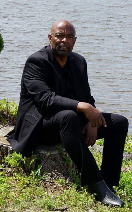 Sheldon Price in a black suit, seated on rocks by the water